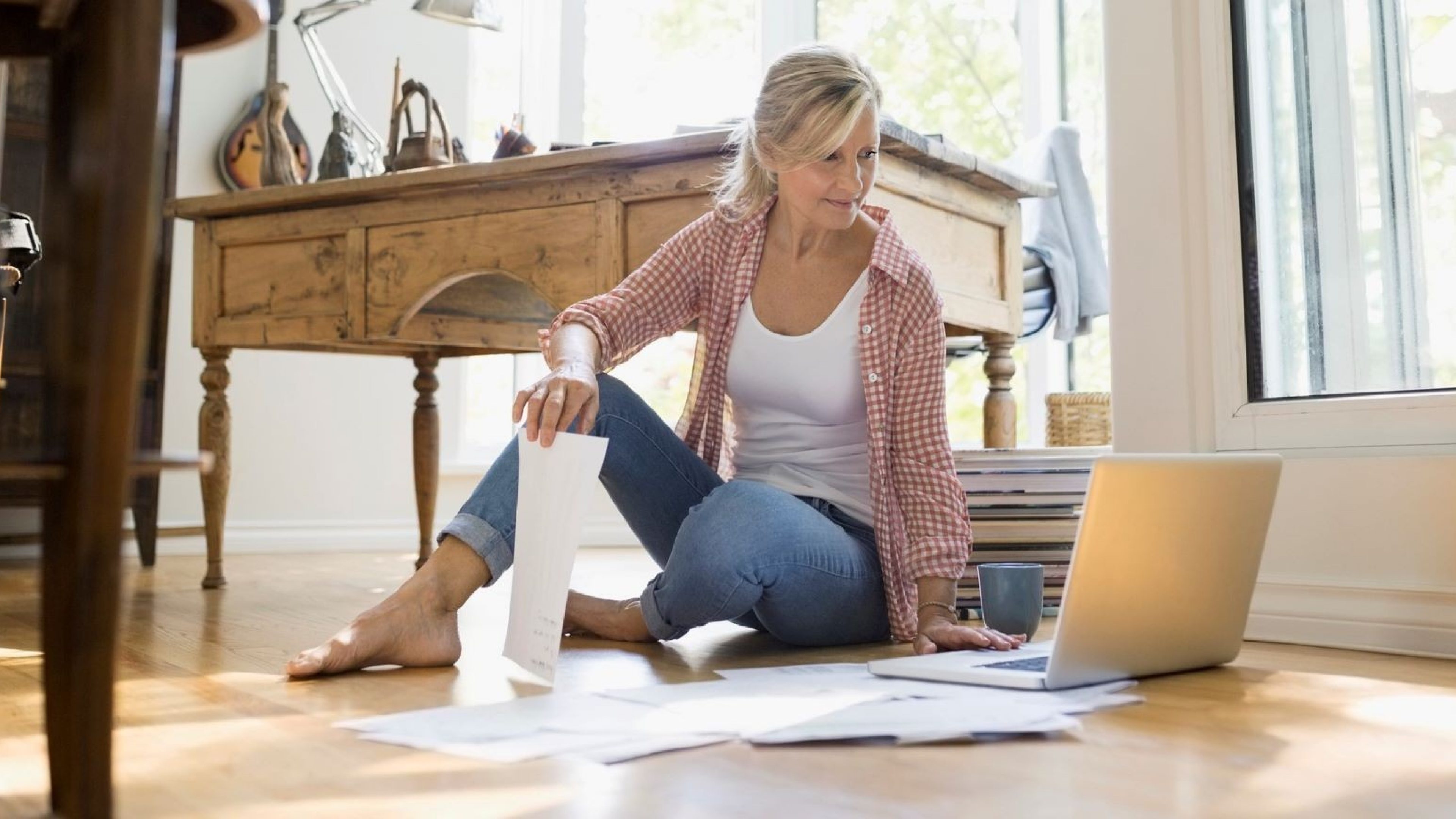 Eine ältere Frau sitzt auf dem Boden mit Papieren und Laptop und prüft die Tragbarkeit ihrer Hypothek im Alter. 