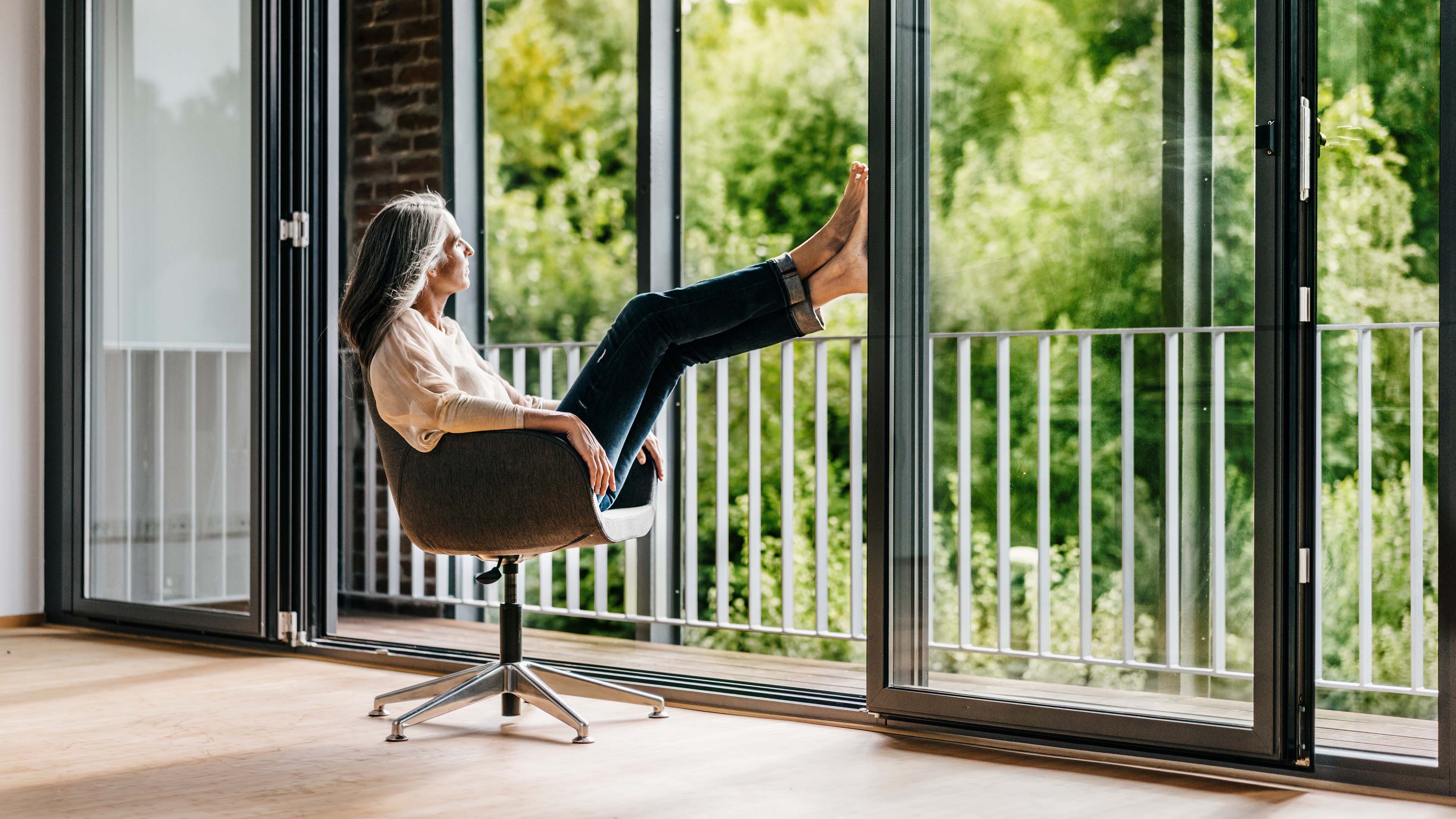 Frau sitzt gemütlich im Sessel und blickt aus einer grossen Fensterfront in die Natur.