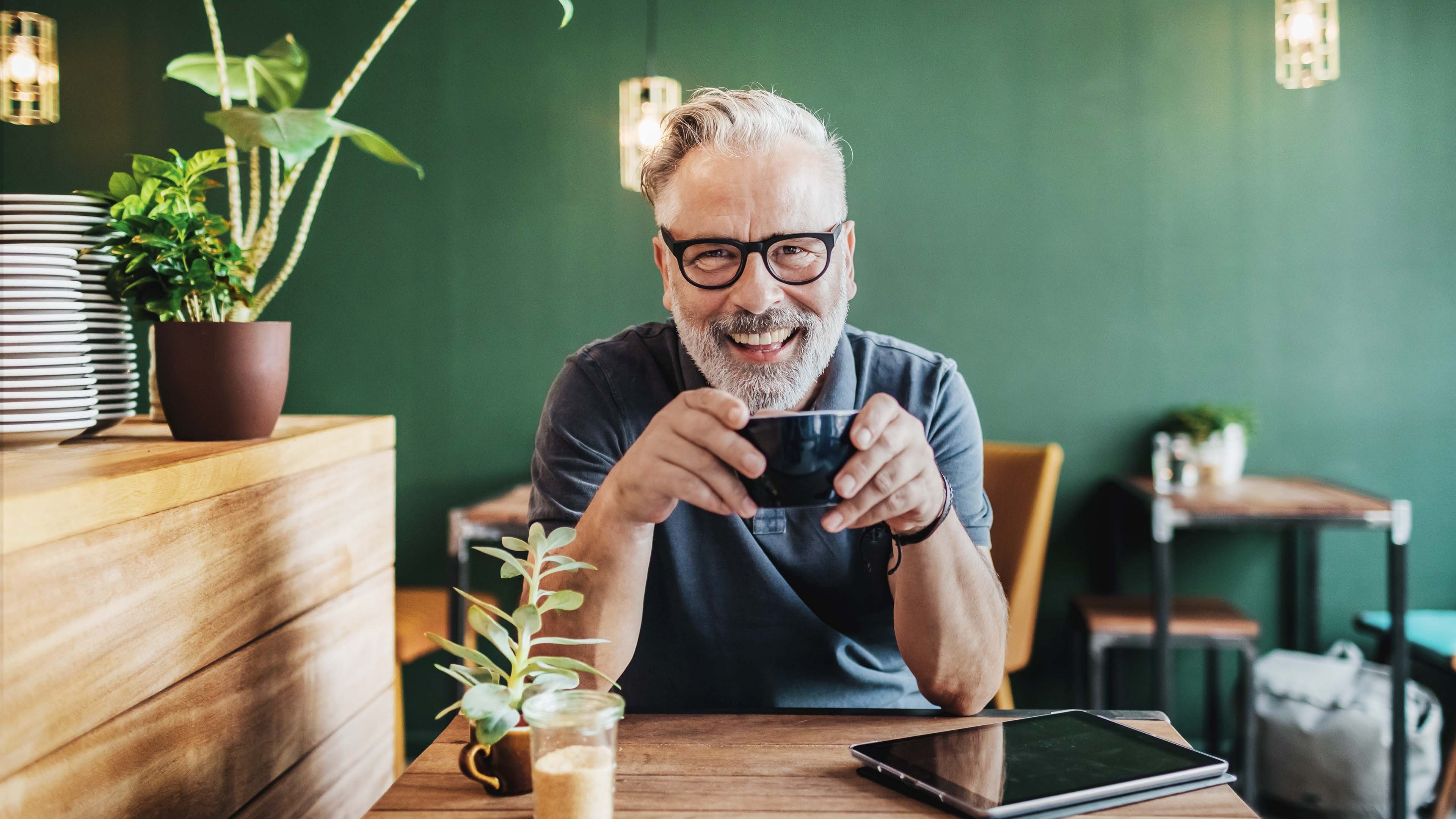 Mann sitzt im Kaffee mit einer Kaffeetasse in der Hand und lächelt in die Kamera